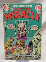 Front cover of Mister Miracle No. 10 comic, showing the prominent metallic head character in a containment device and a group of people in yellow hazmat suits working on some equipment.