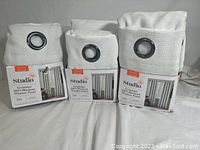 Three white linen-textured blackout curtain panels folded with black metal grommets, packaged with product labels showing brand Studio 3B and product details.