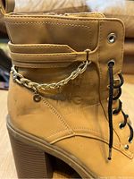 Close side angle photo showing detailed gold chain and black lace-up front on tan heeled boot with fuzzy white tongue