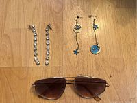 Two pairs of earrings shown arranged above a pair of brown tint aviator sunglasses on wooden surface.
