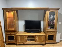 Front view of large wooden entertainment center with TV placed in center space, two vertical glass door cabinets on each side, wooden paneling, and base storage units.