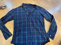 Blue and green plaid flannel shirt laid flat on floor, showing front button-up and long sleeves