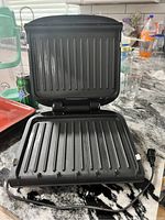 Open top and bottom grill plates of the George Foreman panini press showing ribbed grilling surfaces and grease catcher
