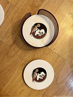 Two round ceramic coasters with a stylized man's face graphic in brown, placed inside and beside a brown metal mesh coaster holder on wood floor.