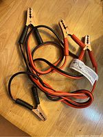 Photo of car jumper cables showing metal clamps with red and black insulated grips and coiled cables on a wooden floor.