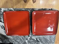 Two red square plates placed side by side on a granite countertop, showing glossy red glaze with minor surface crazing.