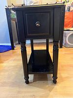 Front view of black wooden side table showing single small drawer with round knob and four turned legs.