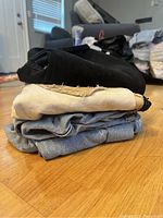 Stack of four pants showing different fabric colors and styles including black, beige with frayed hems, and denim shades.