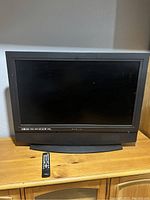 Black Sylvania flat screen TV shown on wooden surface with remote in front, condition worn with scratches.