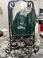 Metal two bottle wine holder with scrollwork, standing upright on kitchen counter, showing front and side views with a green reusable bag in background.