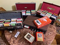 Overall view of 8 track player on table surrounded by multiple cases holding 8 track tapes, a box for a cassette adapter, and loose 8 track tape cases on the table.