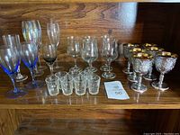 Photo showing 25 assorted glasses including blue-stemmed wine glasses, tall clear flutes, medium clear wine glasses, shot glasses, and ornate silver-tone goblets with gold interiors.