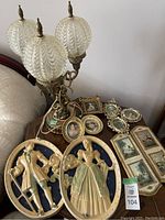 Overview of romantic decorative lot including two large porcelain plaques, small framed miniatures, lamp, and hinged panel on table next to upholstered chair.