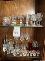 Various glassware including cut crystal champagne coupes, wine glasses, water goblets, shot glasses, small glass bells, and red glass votive holders on wooden shelves.