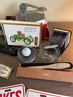 Photo showing multiple vintage office items including mechanical calculator, hole punch, Zippo letter opener, golf ball with stand, glass ashtrays, and partial view of license plate sayings.