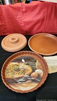 Photo showing three Copco Country Clay non-stick bakeware items: a quiche pan with label, a round pizza pan, and a covered baking dish with lid in natural terracotta color.