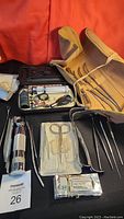 Photo showing assorted metal and plastic medical tools including otoscope, scissors, forceps, probes, and otoscope tips in a beige roll-up case on a black fabric background.