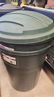 Green Rubbermaid trash can with lid on top, showing general shape and condition.