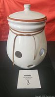 Onion keeper with lid on top, showing curved white ceramic body with ventilation holes and decorative string tied around the neck.