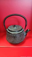 Side angle of antique Japanese iron teapot showing textured ribbed surface and lid design with curved handle on red background.
