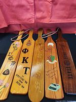 Four traditional double-sided college paddles laid side-by-side, showing painted and etched designs, Greek letters, crests, and personal inscriptions.