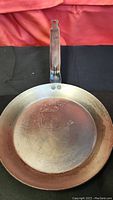 Full view of round copper frying pan with metal handle against red and black background.