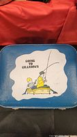 Top view of the small blue suitcase featuring the illustration of two boys fishing and the text "GOING TO GRANDPA'S." The suitcase surface appears worn with scuff marks and nicks.