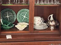 Cabbage leaf dinner plates, lunch plates, cups, serving dishes, and trivet inside wooden cabinet.