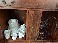 Porcelain china cups, saucers, plates stacked inside a wooden cabinet with glass serving dishes on the side.
