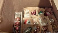 Box of vintage Christmas baubles in multiple compartments separated by cardboard dividers; assorted colors and styles visible inside, some wrapped in tissue paper.