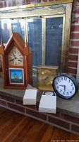 Lot with all clocks shown together on fireplace hearth: wooden pendulum clock with ship picture, brass Angelus quartz clock, three plastic battery clocks, two still in boxes. Background shows fireplace and glass door.