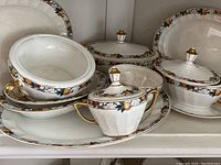 Full lot of Pope Gosser china serving pieces showing bowls, plates, and sugar bowl with lids and handles, all decorated with a fruit and floral motif with gold trim.