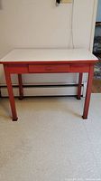 Front view of the vintage enamel top table showing its white top and red painted wooden base with one drawer.