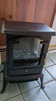 Front and angled views of the black electric infrared fireplace stove heater with metal frame, glass door, and plastic handle.