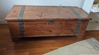 Full view of rectangular wooden cedar chest with metal straps and flat lid on hardwood floor