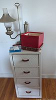 Photo showing white small chest of drawers with basketweave texture and brown bakelite handles, with pewter desk lamp, red box of dolls, blue first aid kit, and silver harmonica on top.