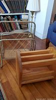 Metal magazine stand with brass-like finish, curved handle, and lattice side panels next to wooden magazine stand on wooden floor.