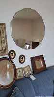 Plain dodecagon mirror with beveled edges hung on wall along with smaller wooden framed mirror, silhouette portraits, oval pressed flower art, and two handwoven pictures on couch below.