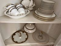 Partial Pavanne china set on shelves, showing stacked dinner plates, salad plates, cups, saucers, and covered serving dish with gold and black trim.