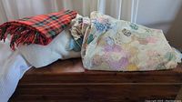 View of the three items on a wooden surface: the plaid throw, cream blanket, and folded pastel patchwork quilt