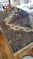 Large Persian rug displayed under dining table with ornate floral borders, cream center field and fringes on two sides. Noted rip on one edge.