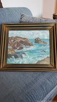 Full view of ocean seascape painting showing waves and rock formations within a wooden frame on a couch.