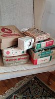 Seven original cardboard boxes of vintage Christmas tree ornaments including Shiny Brite and others stacked on a chair.