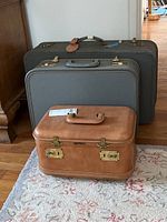 Three vintage suitcases stacked on a floor rug; two large gray suitcases and one smaller brown cosmetic case in front.