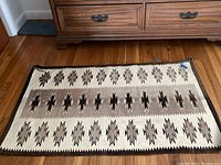 Wool toss rug with Native American motif in black, brown, and cream laid on wood floor in front of drawer chest. Shows overall wear and some edge fraying.
