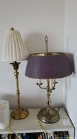 Two satin brass buffet lamps on a white surface, one with white pleated shade the other with purple shade and candlestick design