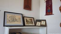 Shelving with several framed pieces of wall art including etchings and prints, vintage wood carving mounted above, and South American woven hanging to right.