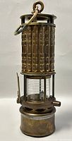 Front view of antique brass miner’s safety lamp showing textured cylindrical body, glass chamber, and base.