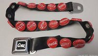 Close-up photo showing the rubber belt decorated with red Coca-Cola bottle caps and a metal buckle labeled 'Coke'.