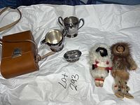 Photo showing Polaroid SX-70 with brown leather case, two Alaska dolls in fur attire, and three silver items including handled cups and a decorative container.